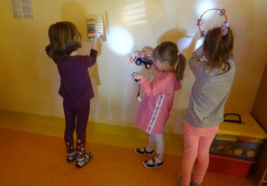 Troje dzieci stoi przy ścianie, trzymają w jednej ręce zabawkę a w drugiej włączone latarki, obserwują powstające cienie na ścianie.